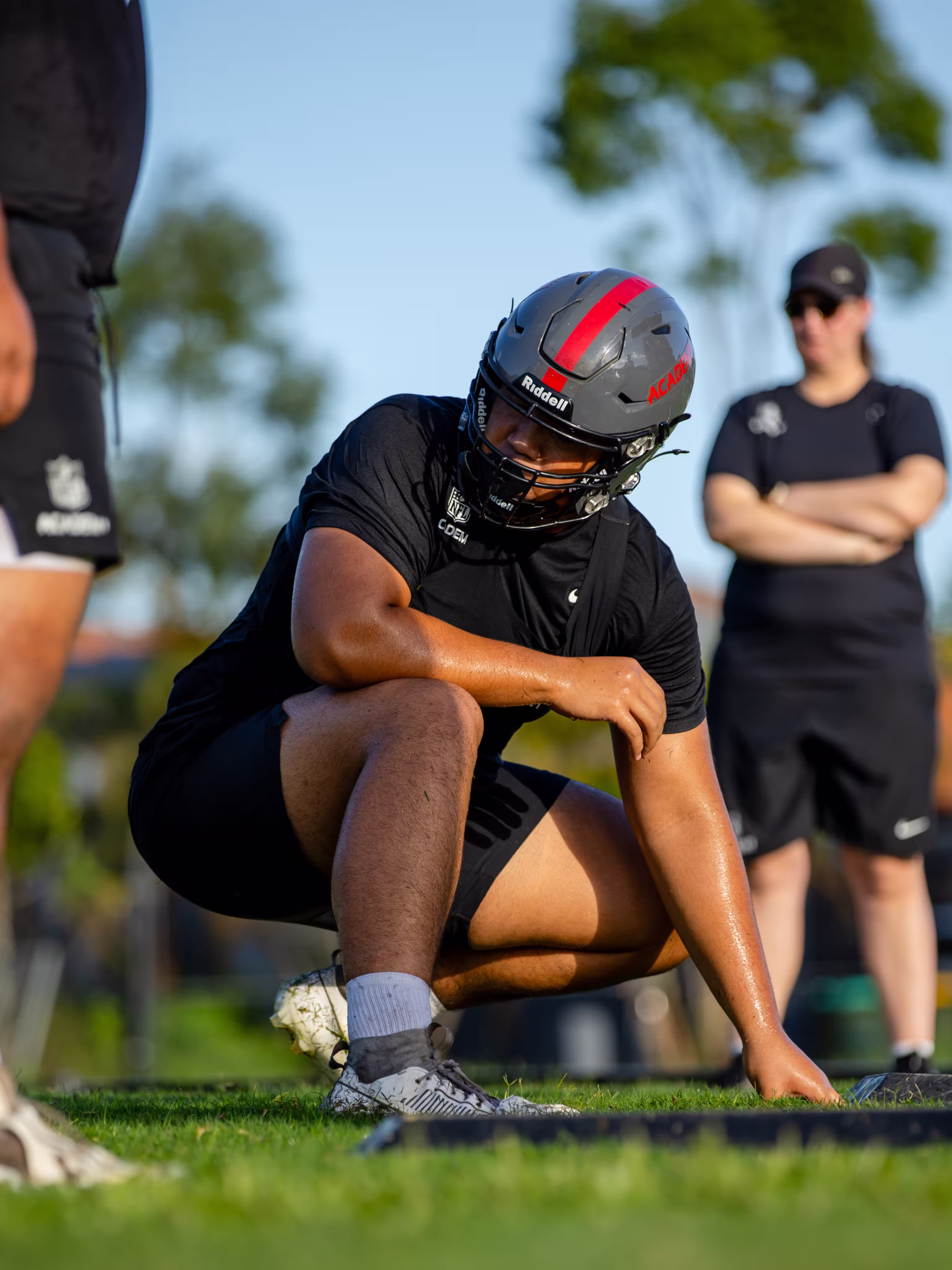 Football player in a helmet crouching on grass during outdoor practice with coach in the background.