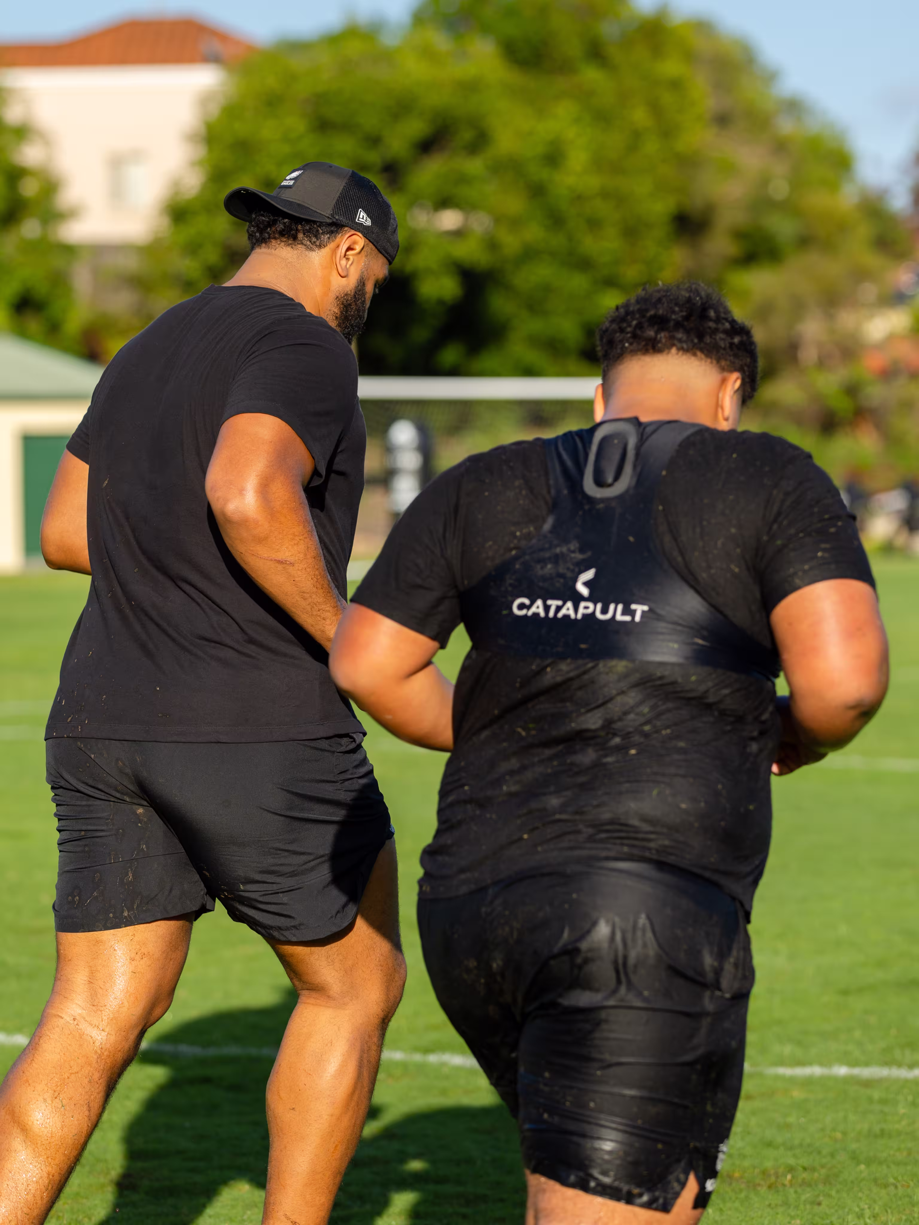 Two men in black sportswear running on grass field, one wearing a Catapult vest for training.