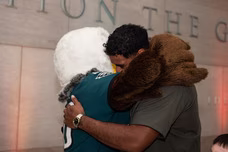 Man in green shirt hugging a person in an eagle mascot costume indoors.