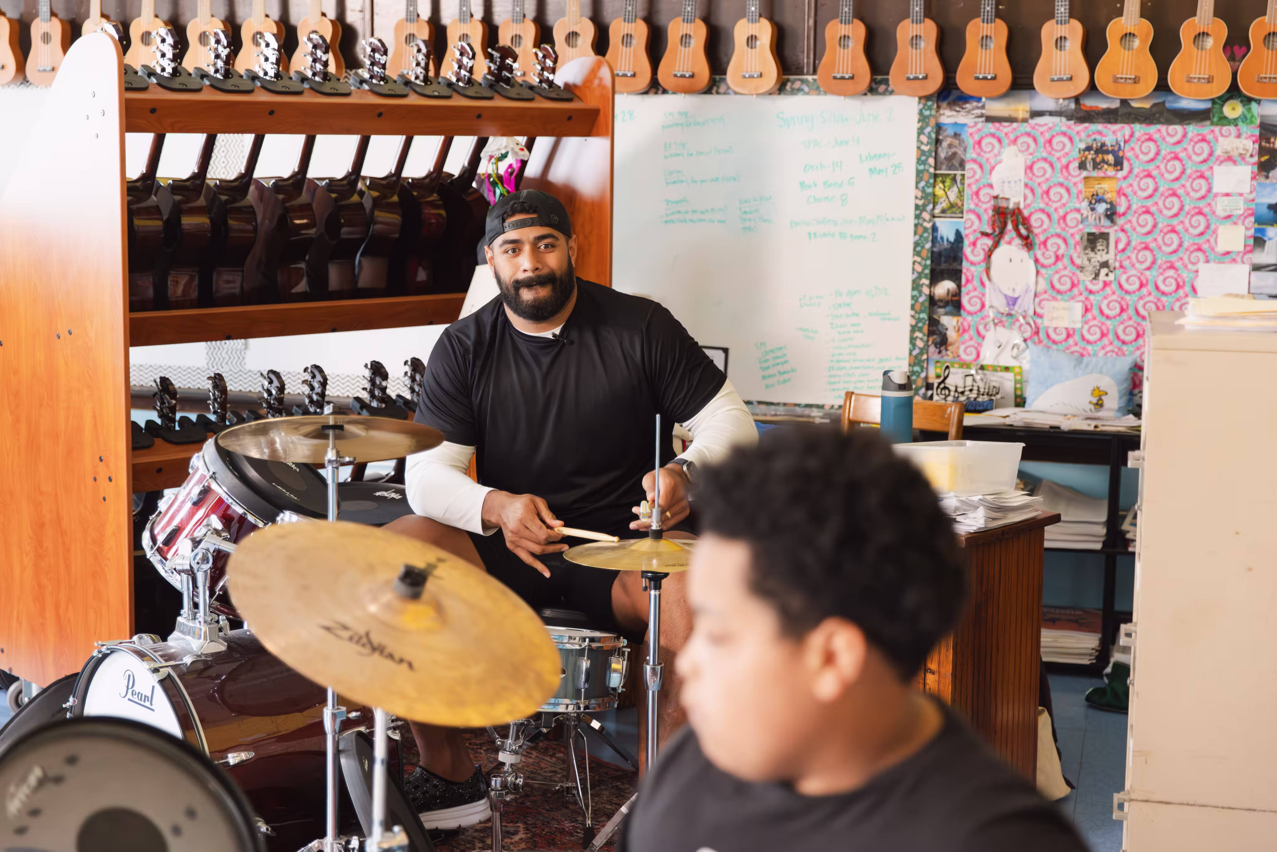 Man in black shirt playing drum set in a music classroom with ukuleles on the wall behind.