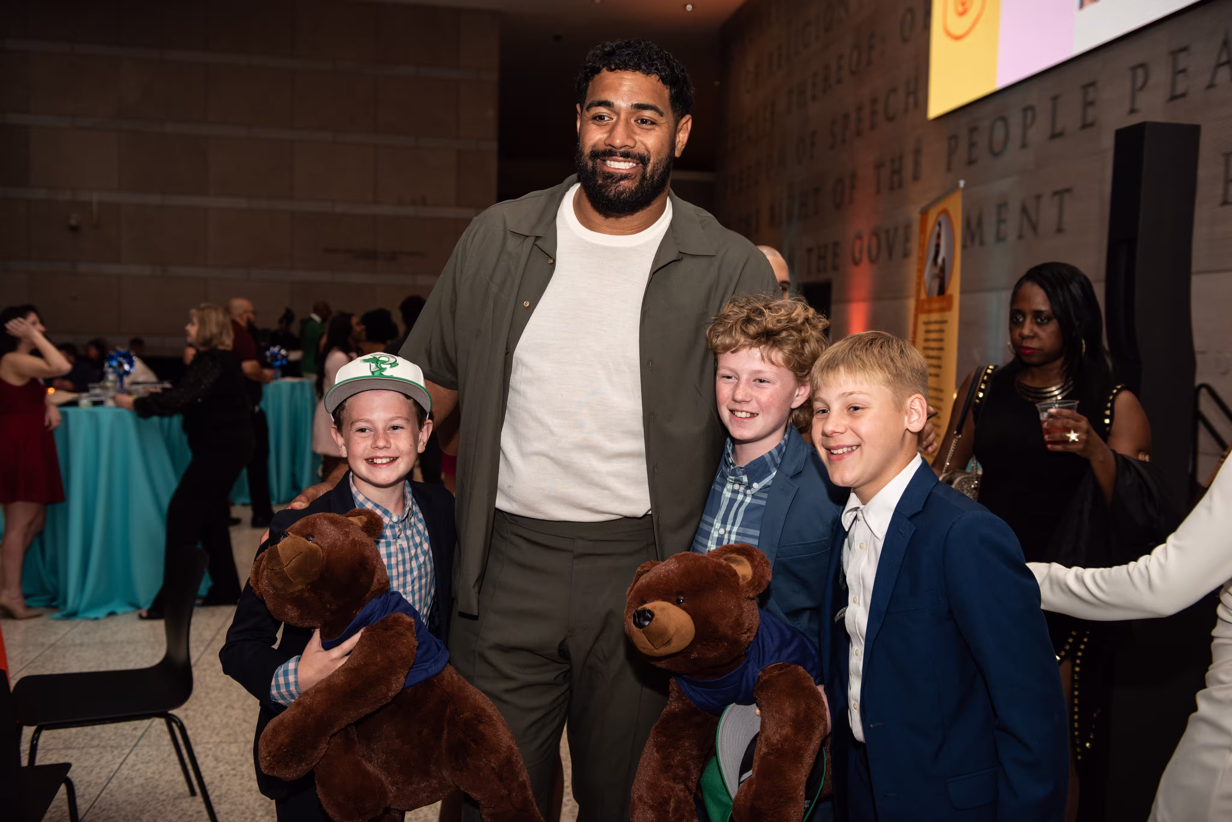 Smiling man hugs three boys holding teddy bears at an indoor event with people in the background.