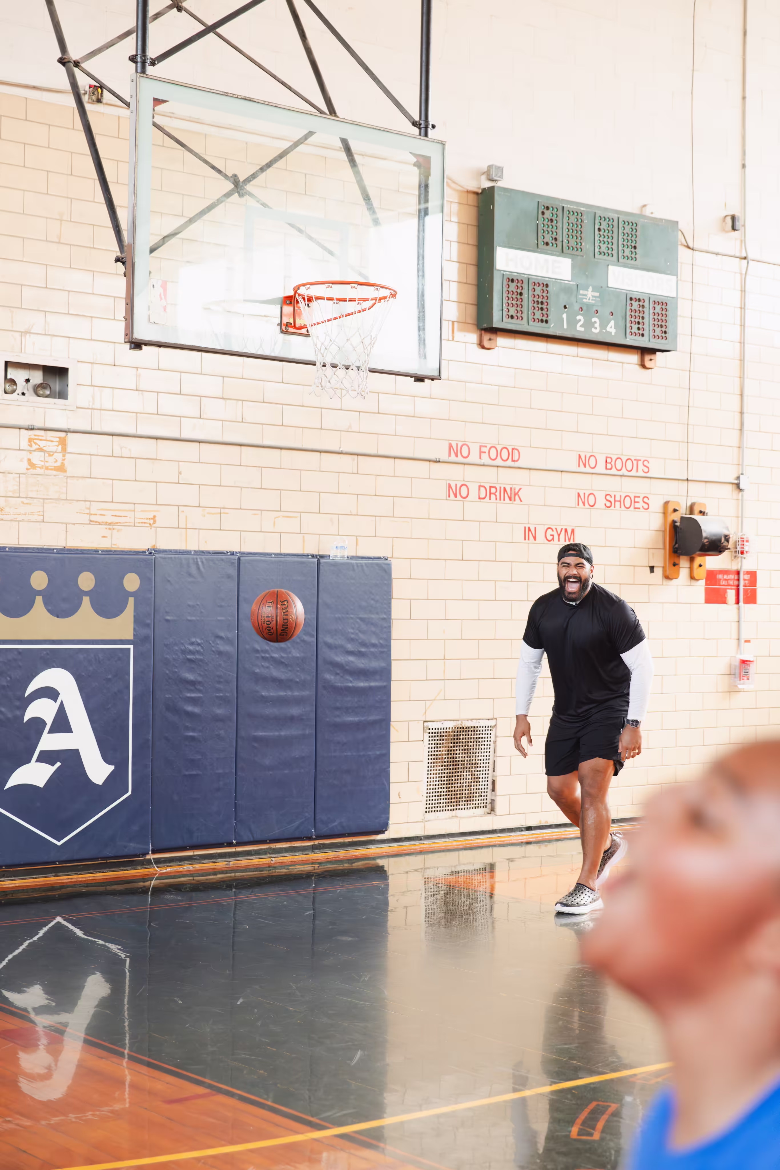 Man in black sportswear smiling in gym with basketball near hoop and scoreboard on wall.