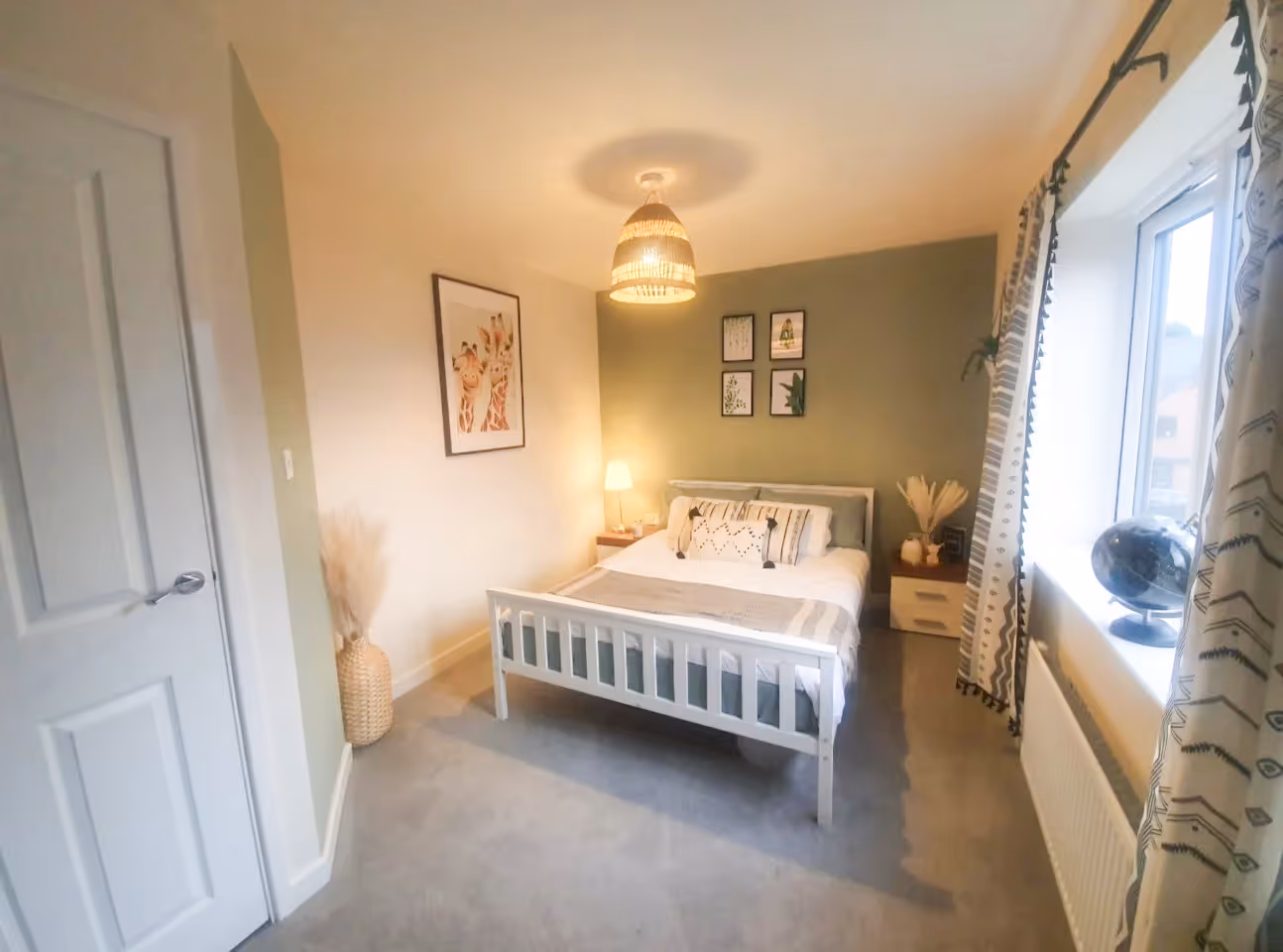 Cozy bedroom with white bed, green accent wall with framed art, and window with patterned curtains.