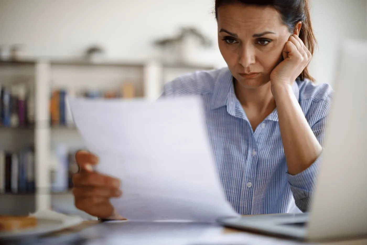 Woman with a concerned expression reads a document while sitting at a desk with a laptop.
