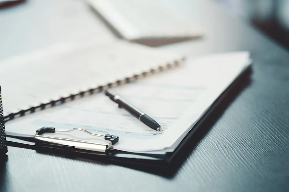 Black pen placed on official documents clipped on a black clipboard on a textured dark surface.