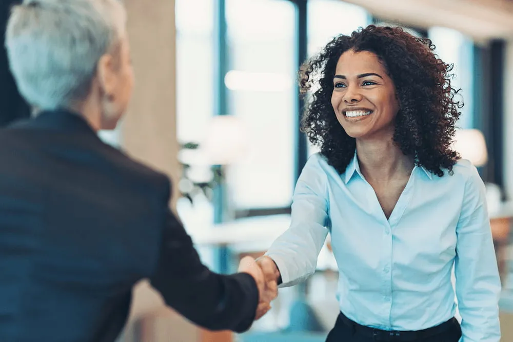 Smiling woman in blue shirt shaking hands with another person in an office setting.