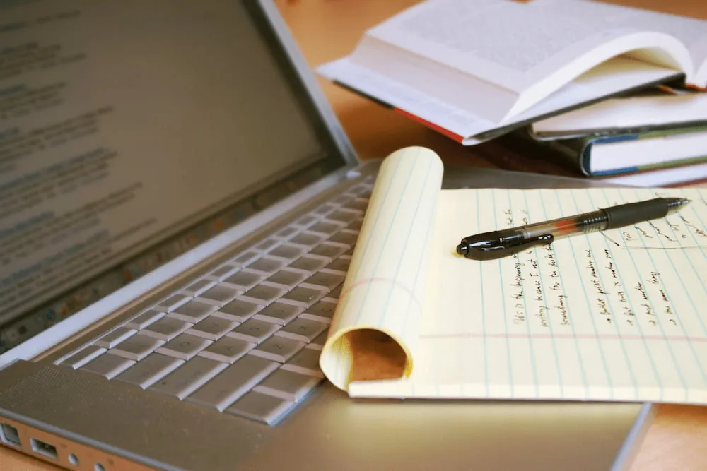 Laptop keyboard with a legal pad and pen on top, and open books in the background on a wooden desk.