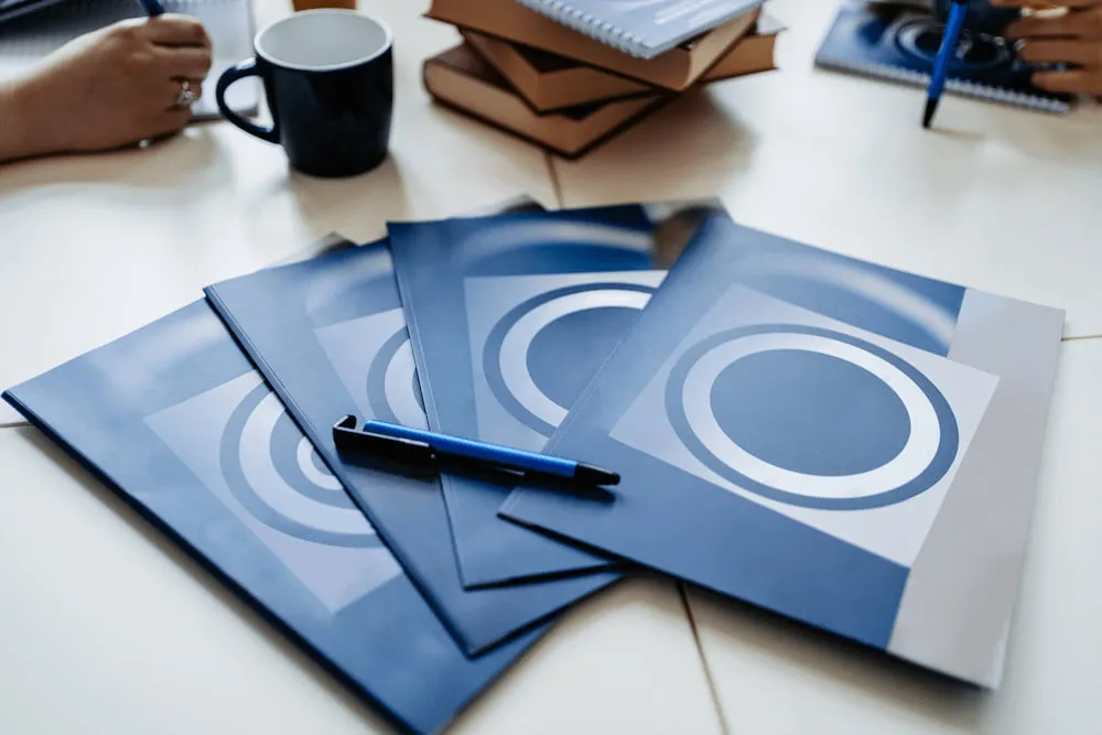 Blue folders with circular logo on a table with a pen, books, coffee cup, and hands holding pens.