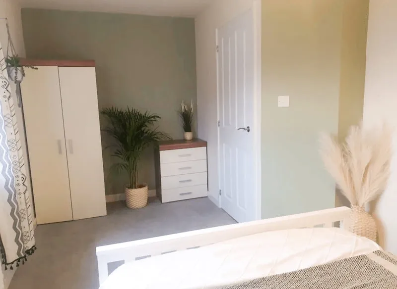 Minimalist bedroom with white wardrobe, chest of drawers, green plant, and cozy bed with patterned blanket.