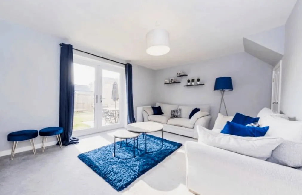 Bright living room with white sofas, blue cushions, rug, stools, floor lamp, and glass door to patio.