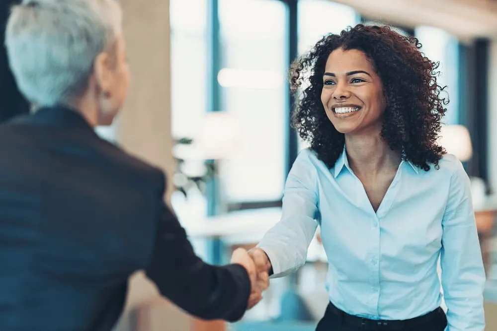 Smiling woman in a blue shirt shaking hands with a person in a black suit in an office.