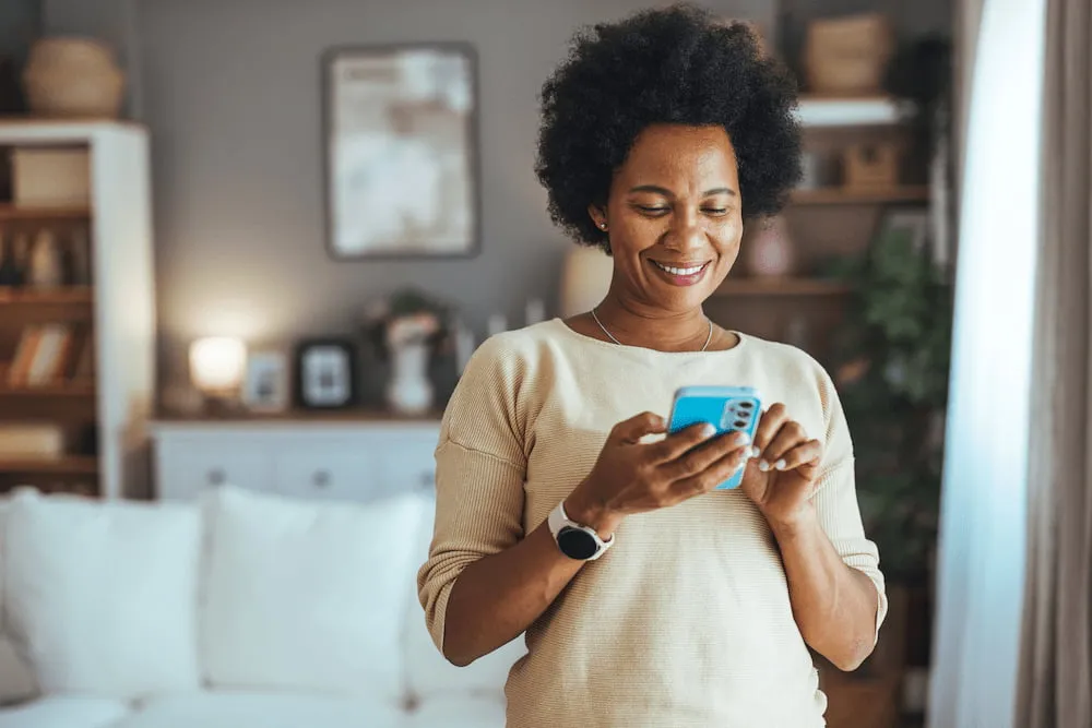 Smiling woman in beige sweater using blue smartphone in a cozy living room.