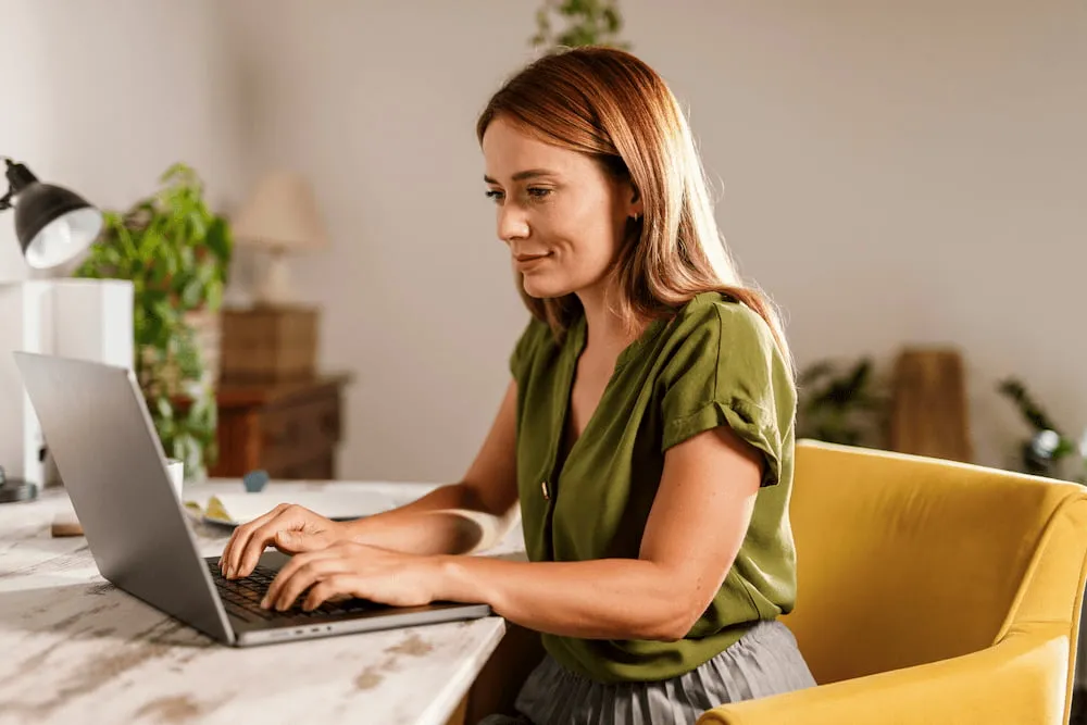 Woman in green blouse typing on laptop while seated in a yellow chair at a desk.