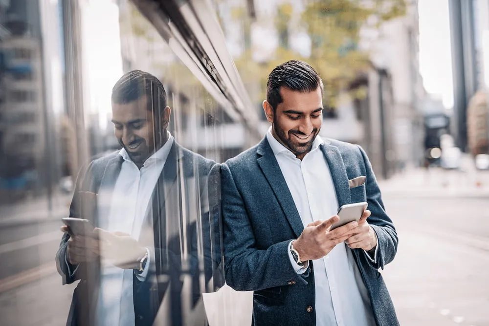 Smiling man in a blazer leaning on glass wall, looking at his phone on a city street.
