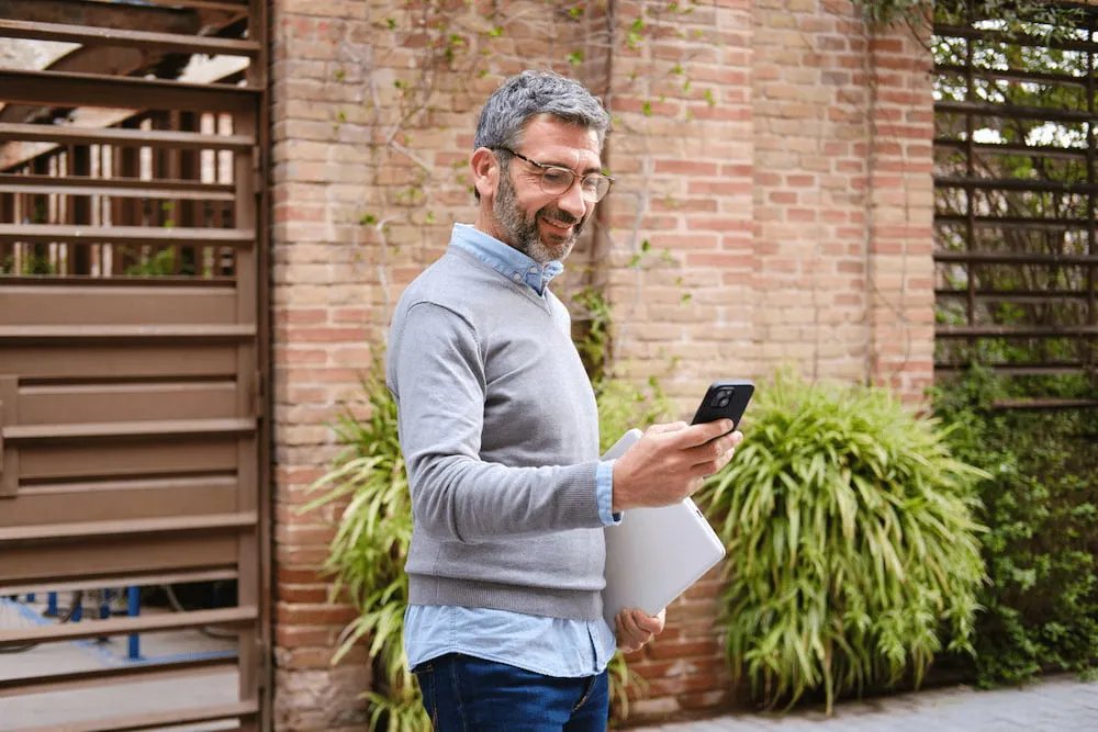 Smiling man with glasses holding laptop and looking at smartphone outdoors near brick wall.
