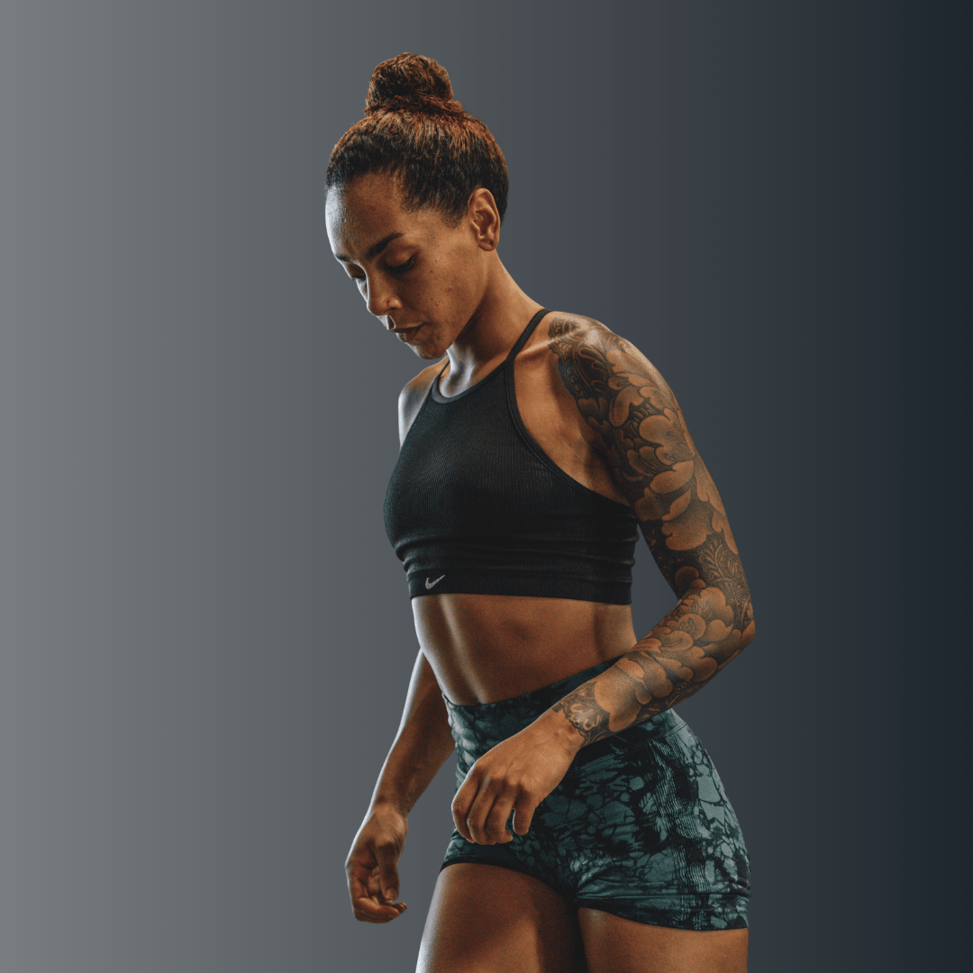 Athletic woman with tattooed arm in black sports bra and patterned shorts looking down against gray background.