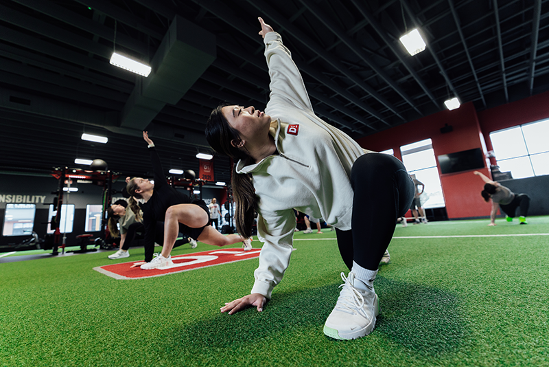 Group in gym stretching on green turf, woman in front reaching upward wearing white sweatshirt and sneakers.
