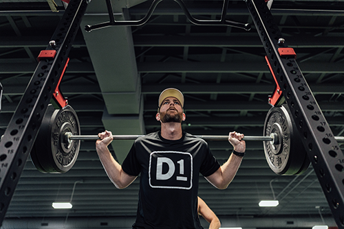 Man wearing a cap and D1 shirt lifting a barbell on a squat rack in a gym.