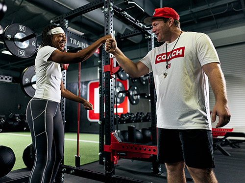 Female athlete and coach fist bump in a gym filled with weights and equipment.