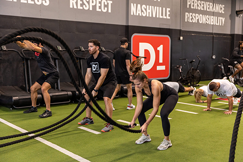 Group fitness class at gym with participants using battle ropes and exercising on turf.