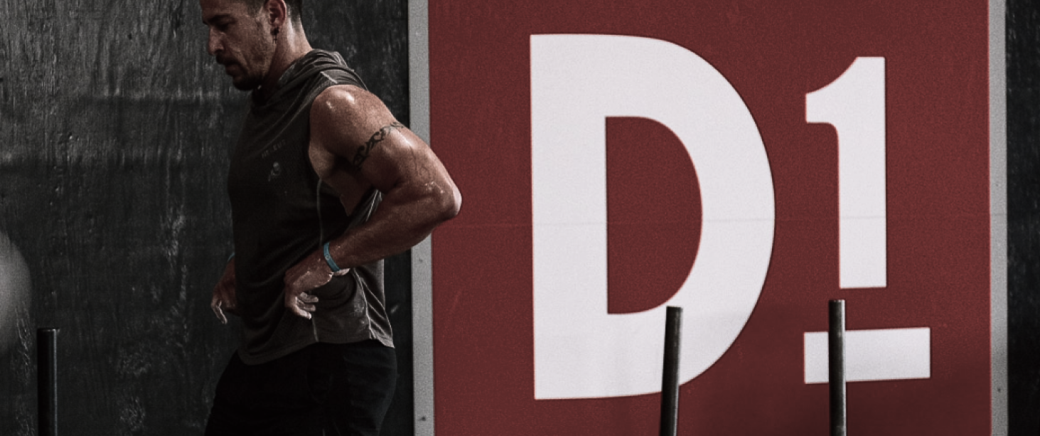 Muscular man in a sleeveless shirt stands by a wall with a large D1 logo during training.