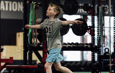 Young boy balancing with arms outstretched inside a gym with weightlifting equipment behind him.