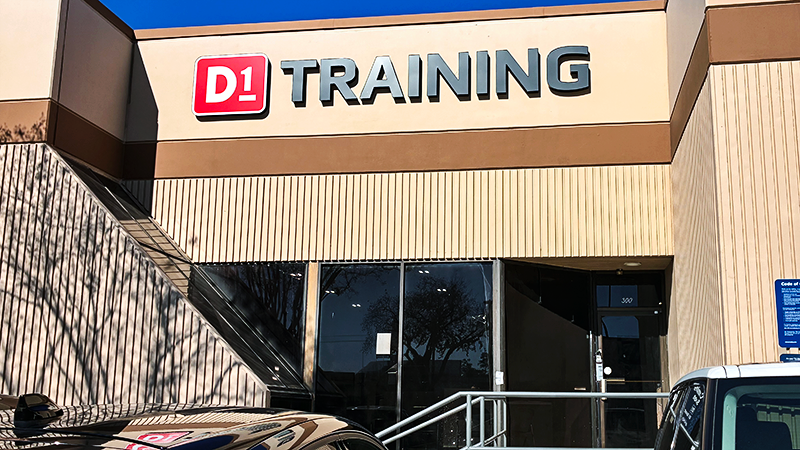Exterior view of a beige building with a D1 Training sign above glass doors and parked cars outside.