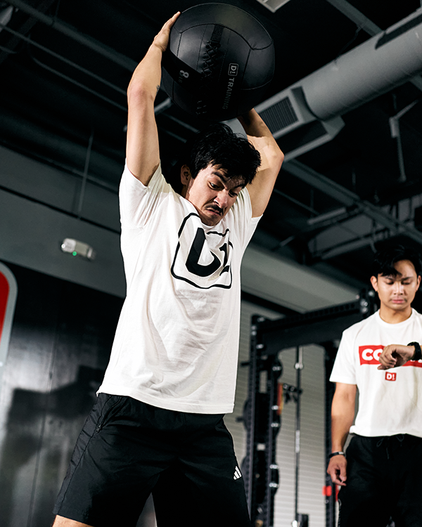 Man lifting a black exercise ball above his head in a gym while another man watches.
