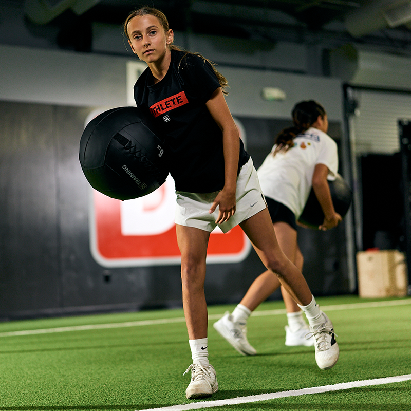 Young athlete in black shirt and white shorts holding a black medicine ball on turf field.