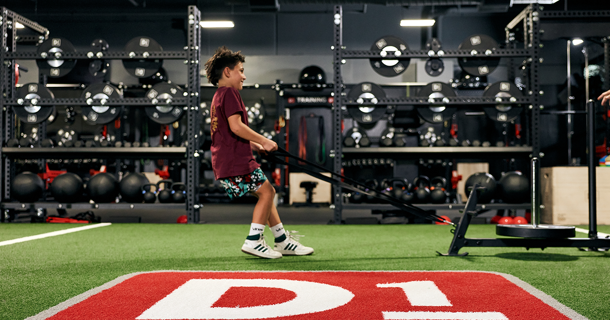 Young boy in gym pulling a weighted sled on artificial turf with fitness equipment in background.