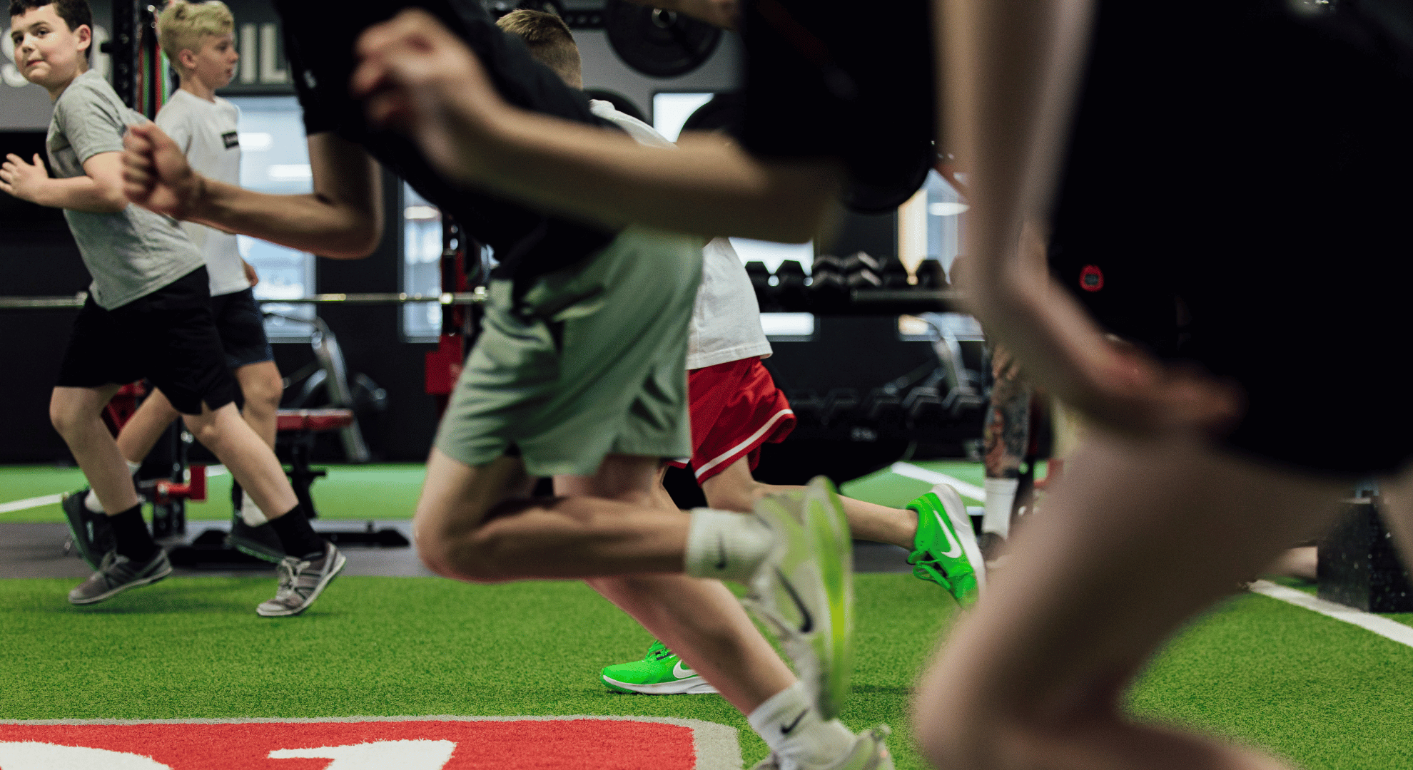 Children running indoors on green turf, with gym equipment in background, in a speed and agility training session.
