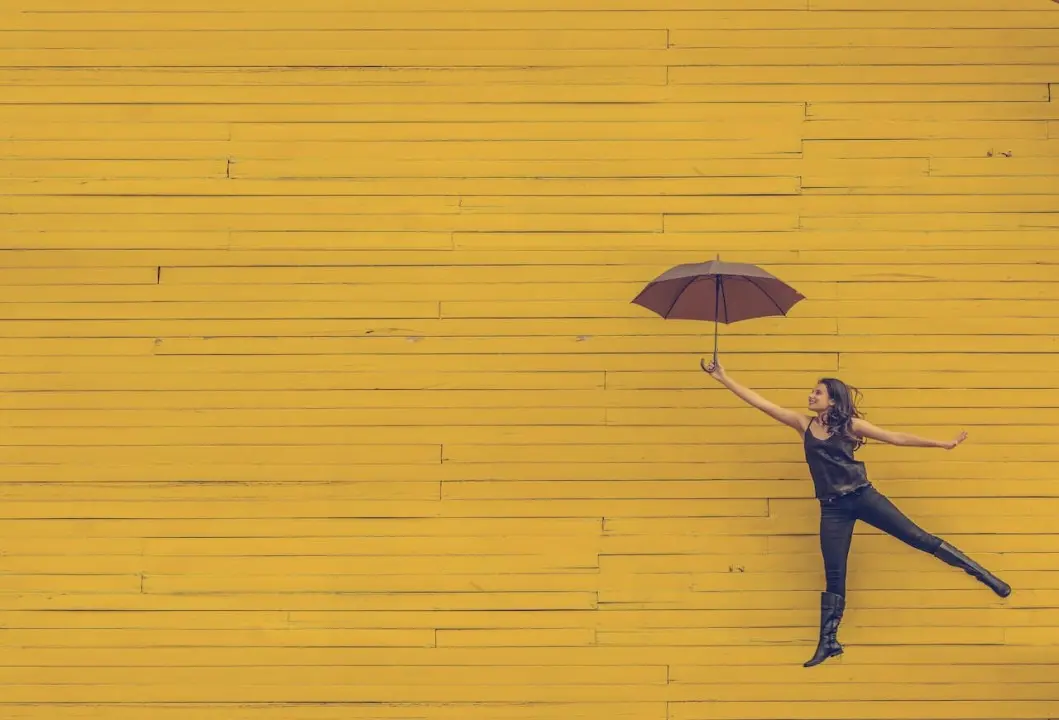 Woman in black outfit holding a brown umbrella, appearing to float against a bright yellow wooden wall.