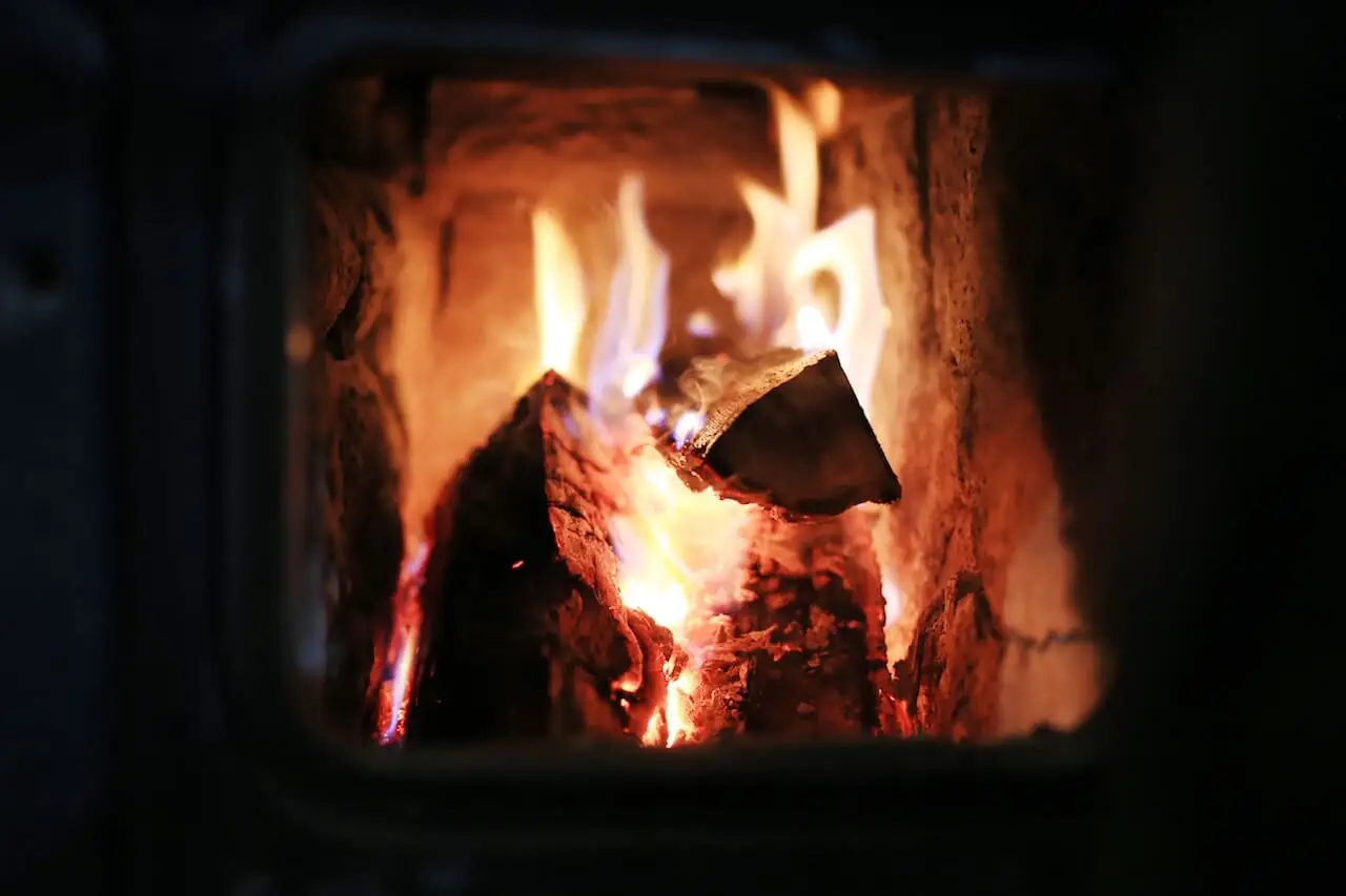 Close-up view of a log fire burning inside a wood stove, with bright orange flames and glowing embers illuminating the brick interior.