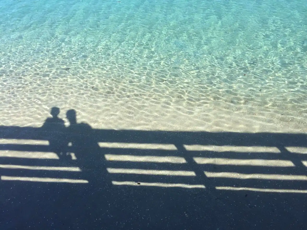 Shadow of an adult and a child leaning on a wooden railing, cast onto clear rippling shallow water.