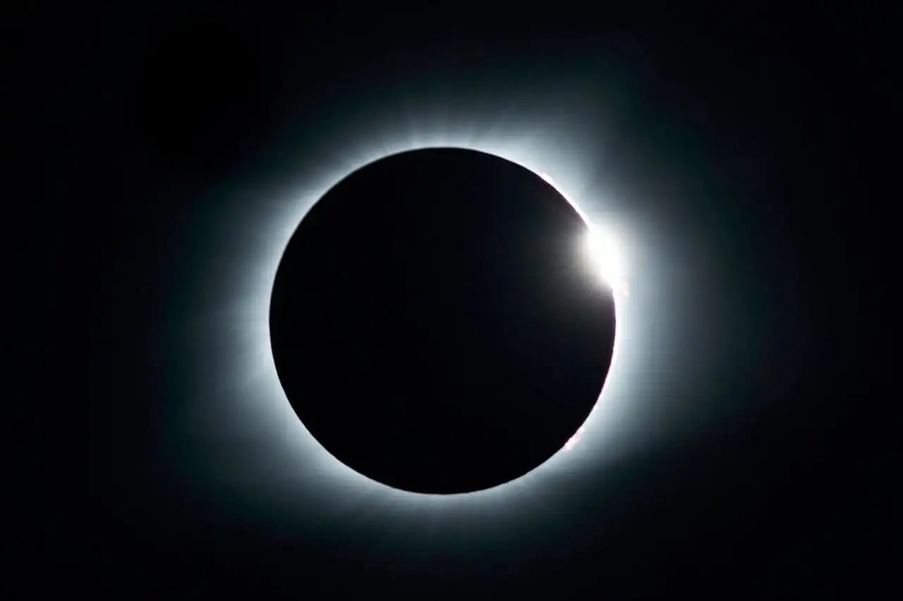 Total solar eclipse with a glowing white corona and a bright burst of light at the edge, set against a dark sky.