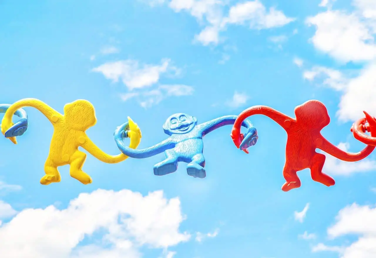 Three colourful plastic monkeys—yellow, blue, and red—linked arm-in-arm against a bright blue sky, symbolising playful yet unbreakable connection, interdependence, and the ties that hold mandalic structures together.