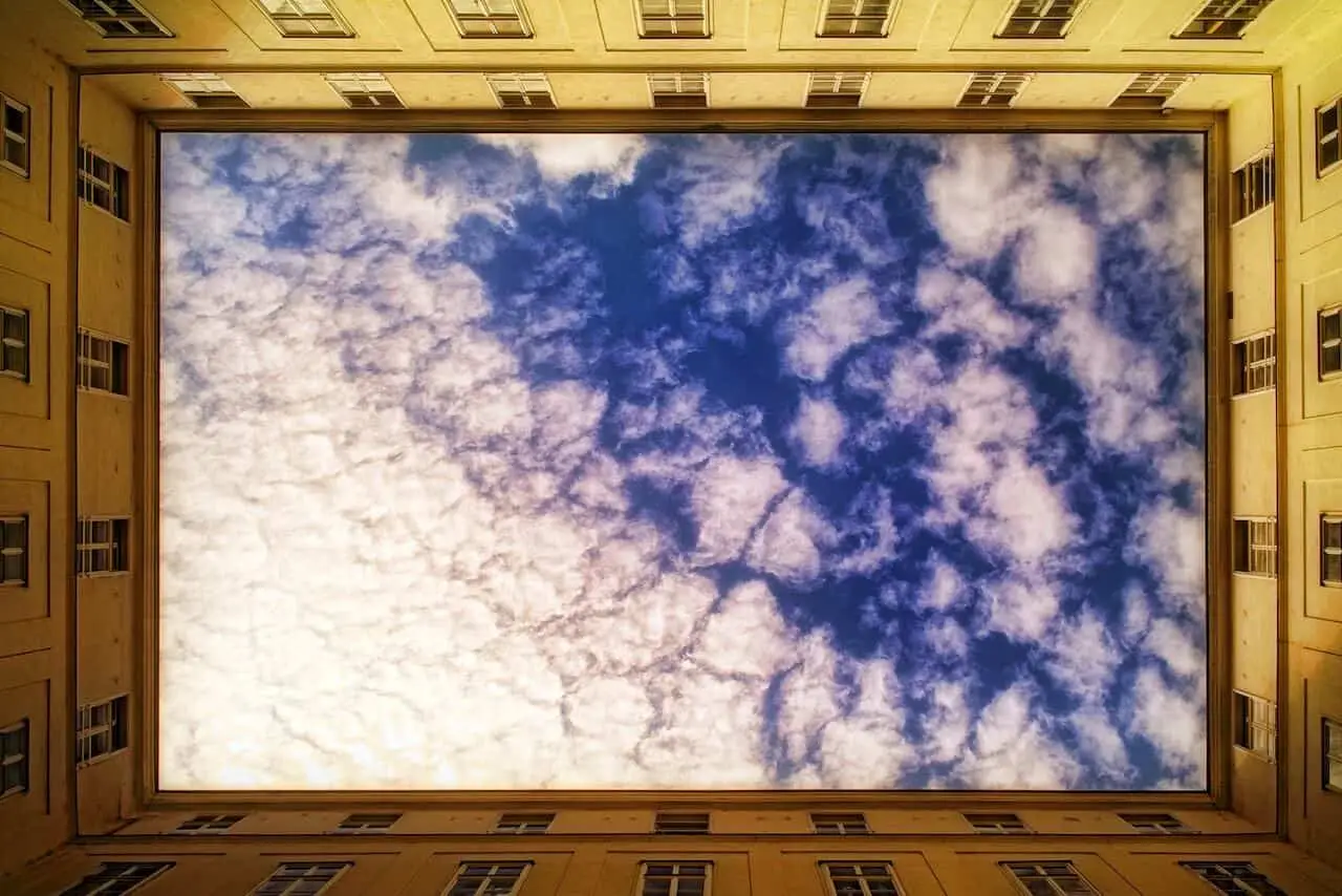 Looking up from within a square courtyard, the building frames a sky filled with textured clouds—symbolising the protective and defining boundary of a mandala.