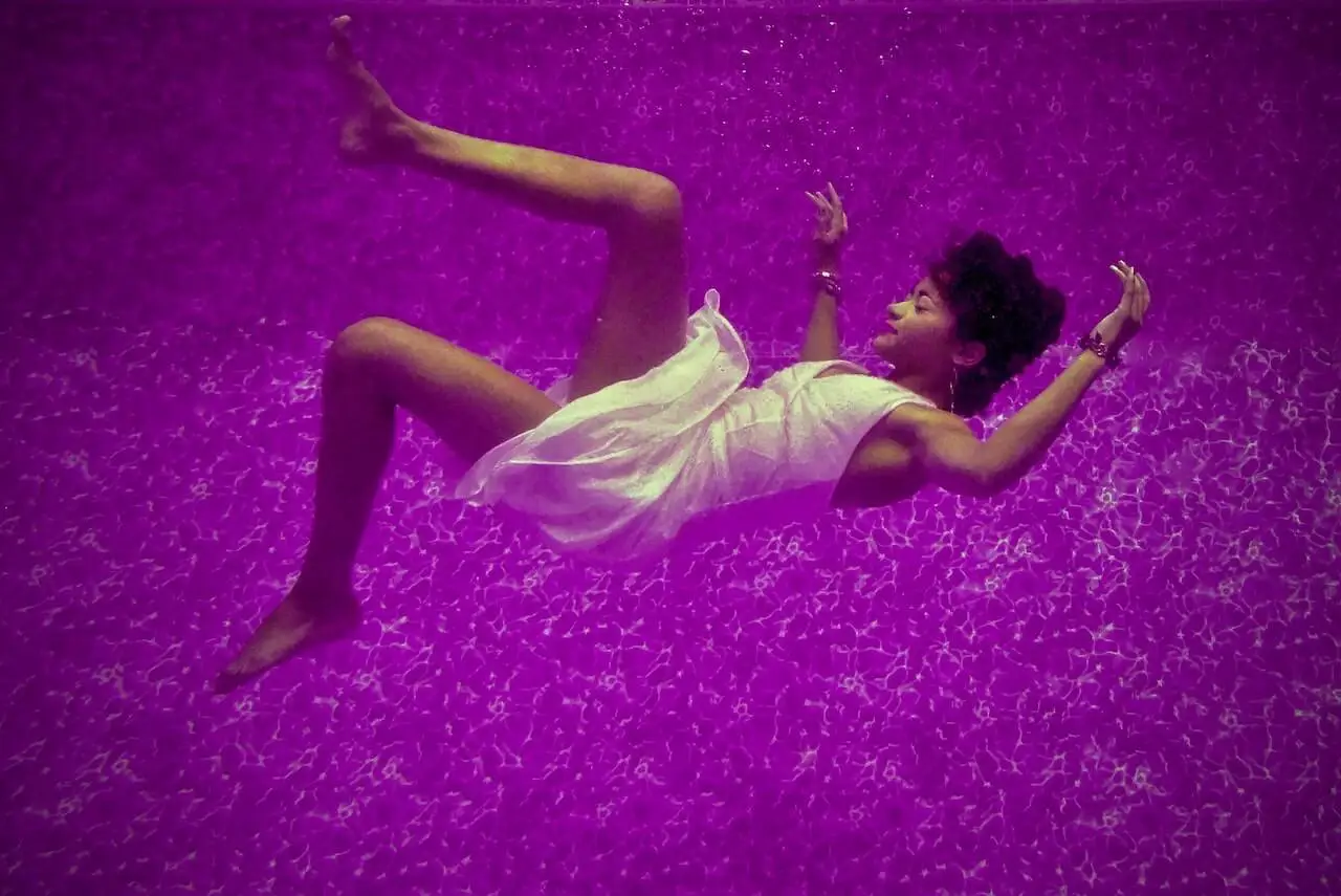 Woman in a white dress floating underwater in a dreamlike pose, surrounded by purple-tinted water and light reflections.