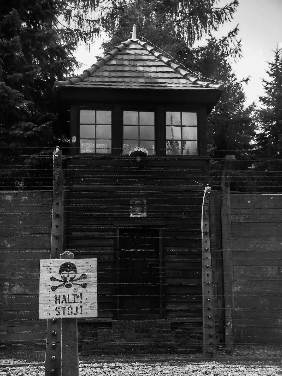 Black and white image of a guard tower and barbed wire fence at a concentration camp, with a warning sign in the foreground reading "HALT! STOJ!" and a skull and crossbones symbol.