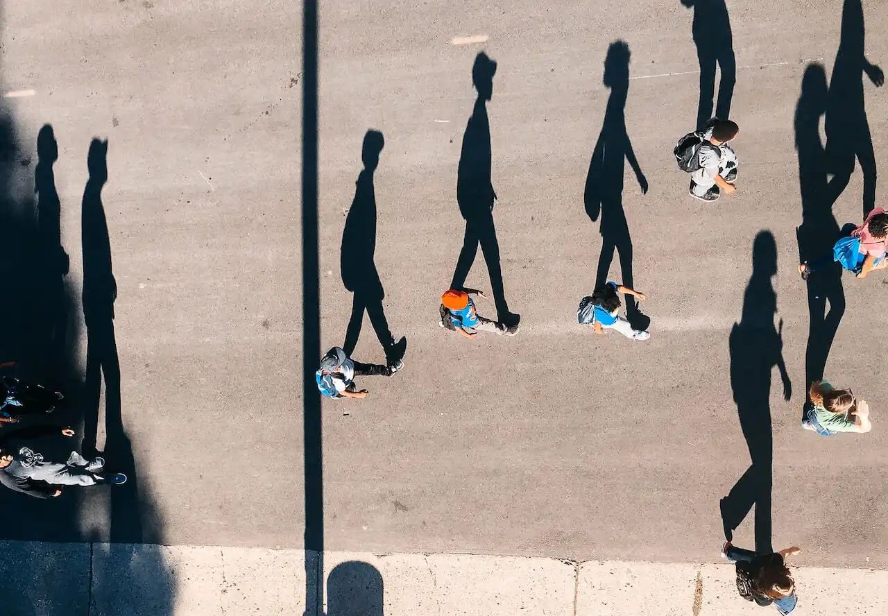 Aerial view of people walking on a paved surface, casting long, distinct shadows in the sunlight. The image captures movement and individuality through shadow play.