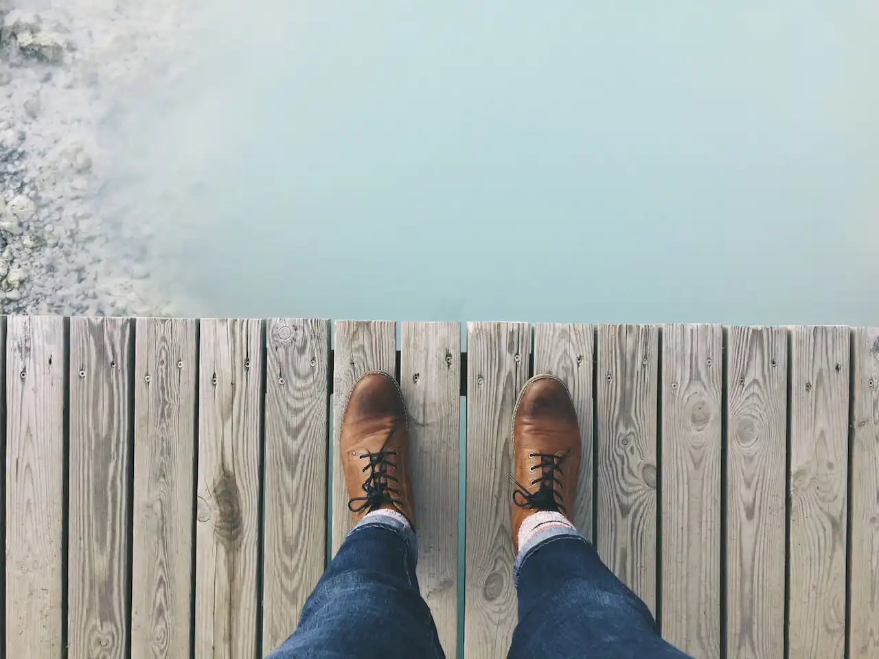 A person's legs and feet in brown leather shoes standing at the edge of a wooden dock, overlooking calm, pale blue water, suggesting a moment of contemplation or transition.