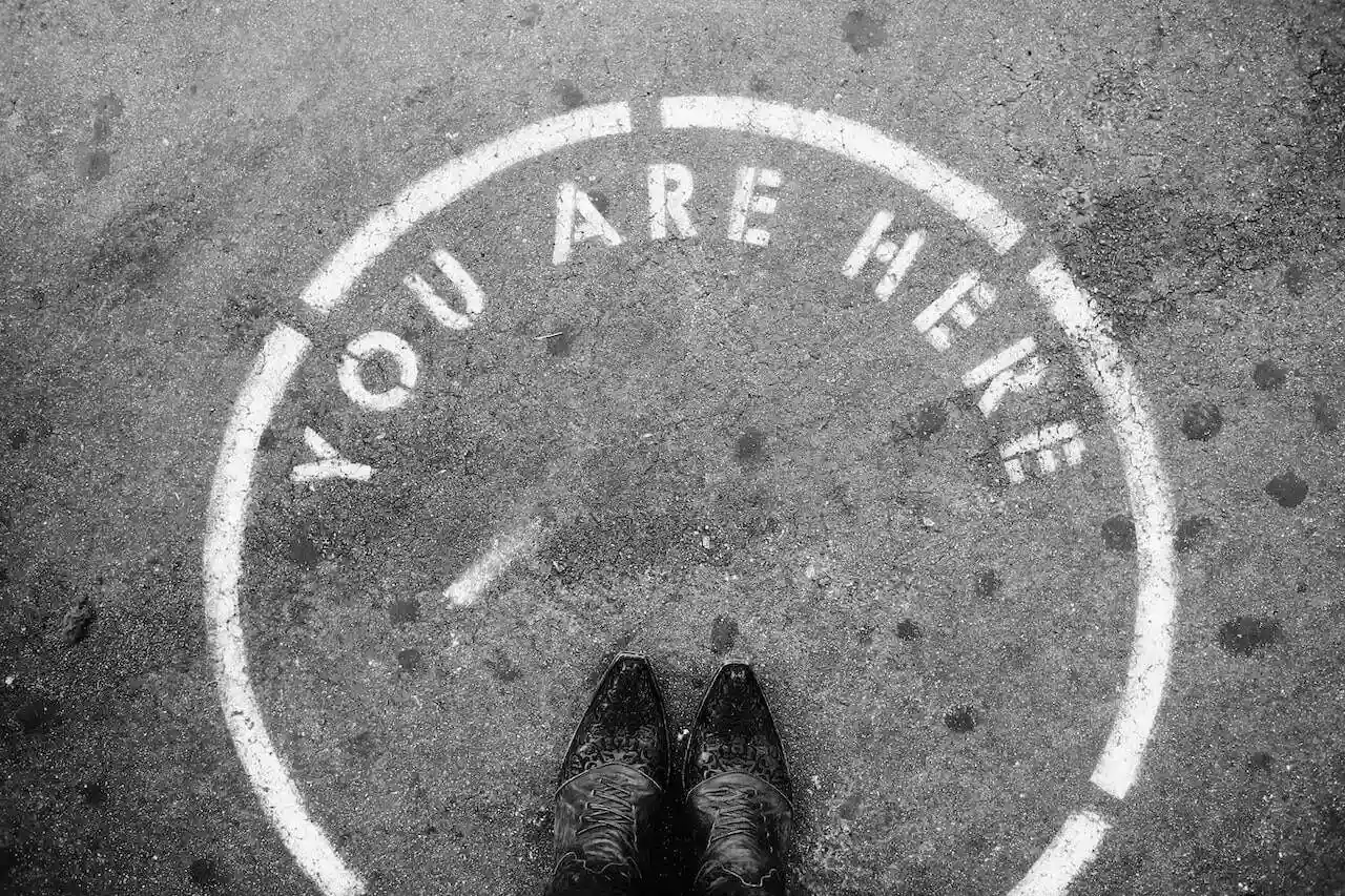 Black-and-white photo showing a pair of shoes standing at the center of a painted circle on the ground that reads "YOU ARE HERE" — symbolising the moment of clarity, acceptance, and presence often needed before meaningful change can begin in organisations.