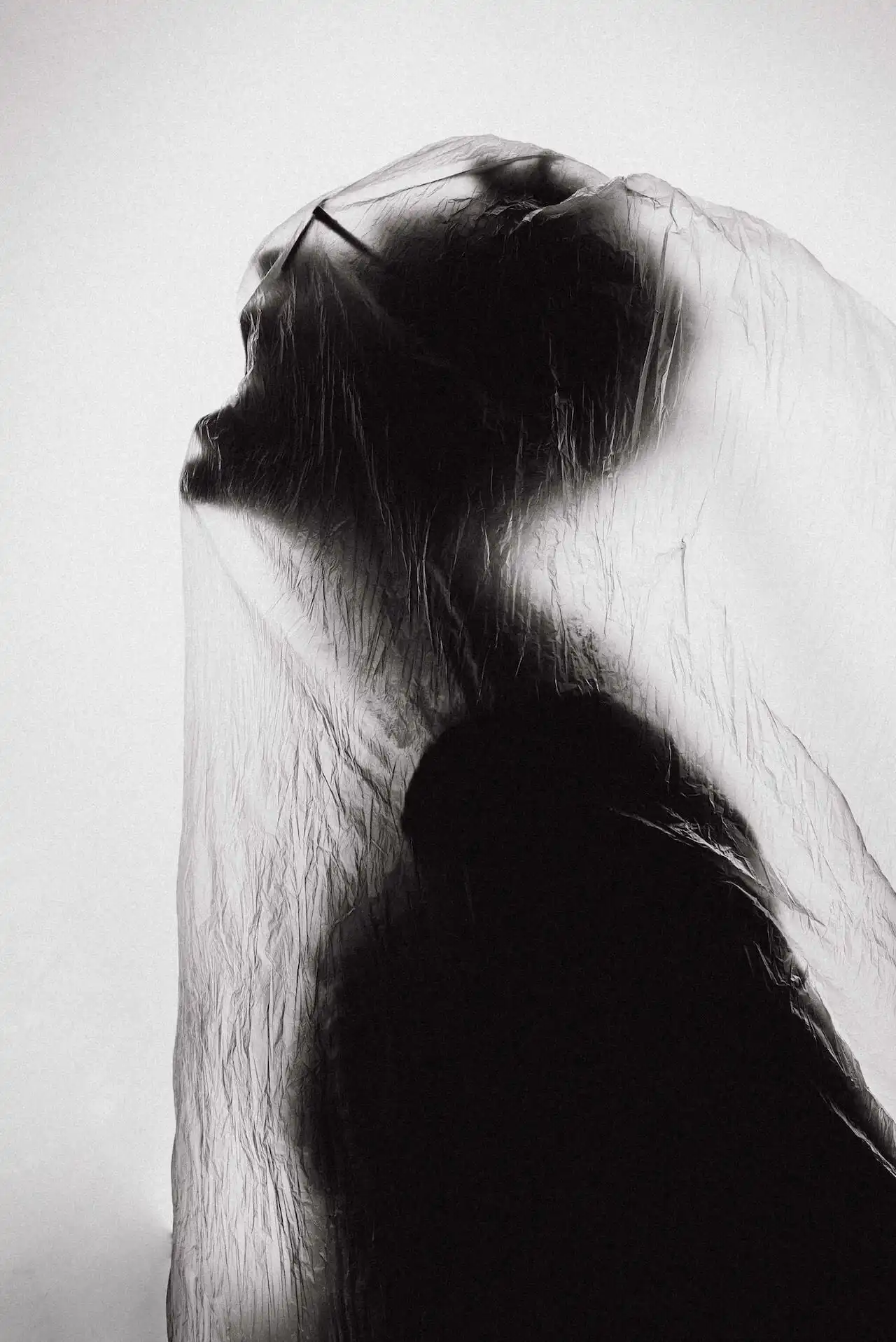 Black and white photo of a person wrapped in translucent plastic, their face contorted in a silent scream, evoking a sense of suffocation, tension, and emotional vulnerability.