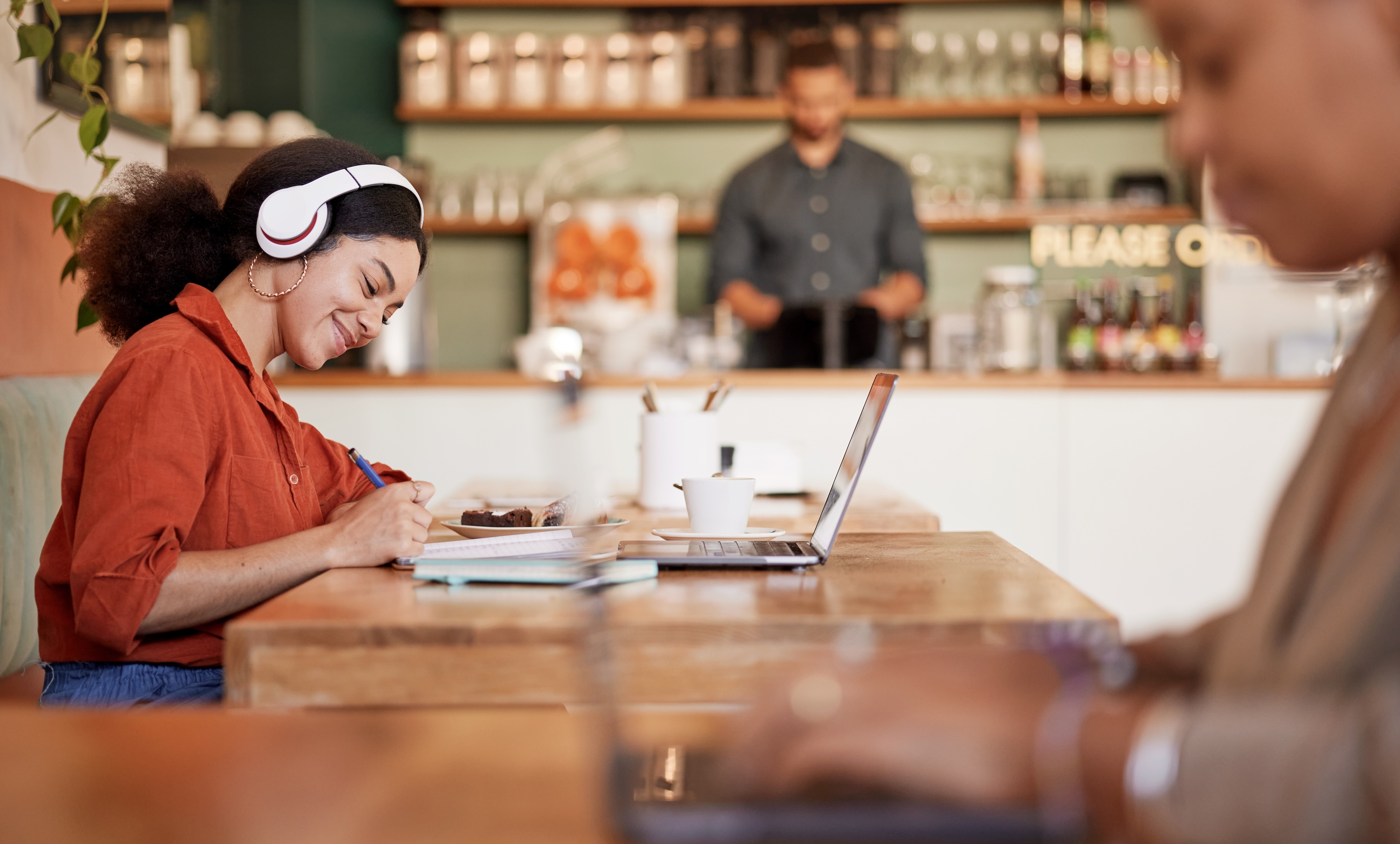 A young woman with headphones on, smiling and studying on her laptop in a coffee shop, representing a convenient work-from-home environment near Altitude a Prosper Community in Houston, TX.