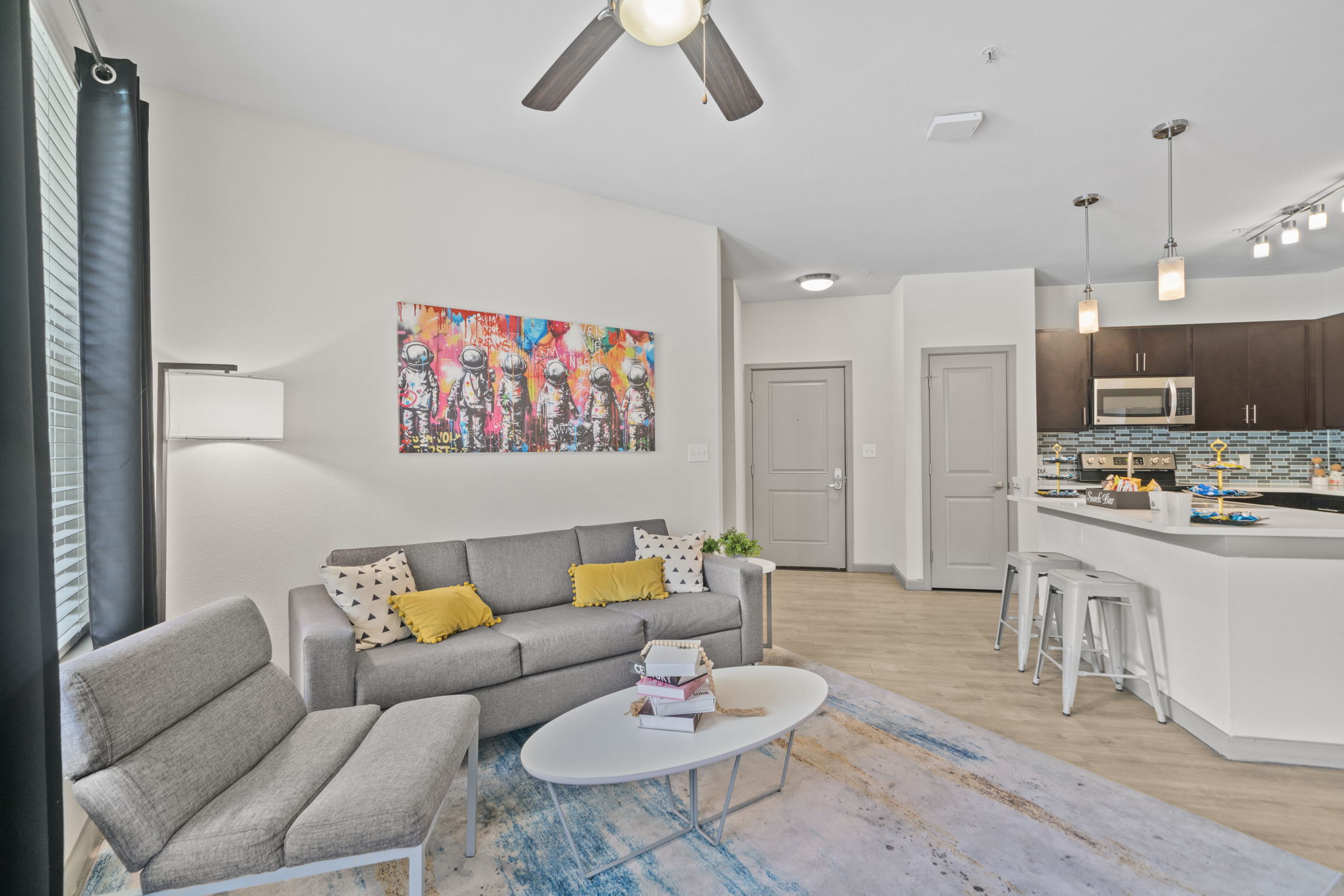 Contemporary living room in an apartment at Altitude in Houston, TX, featuring a grey sofa, colorful art, and an open layout connecting to the kitchen.