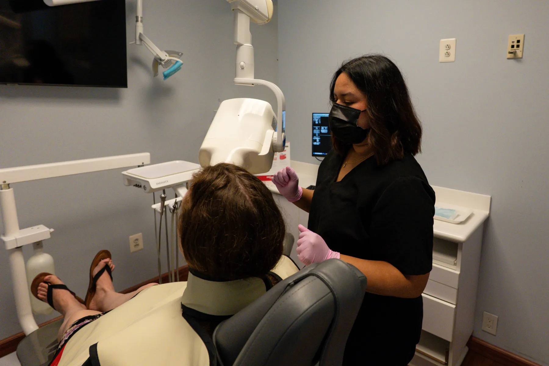 Dentist wearing mask and gloves preparing to examine patient seated in dental chair.