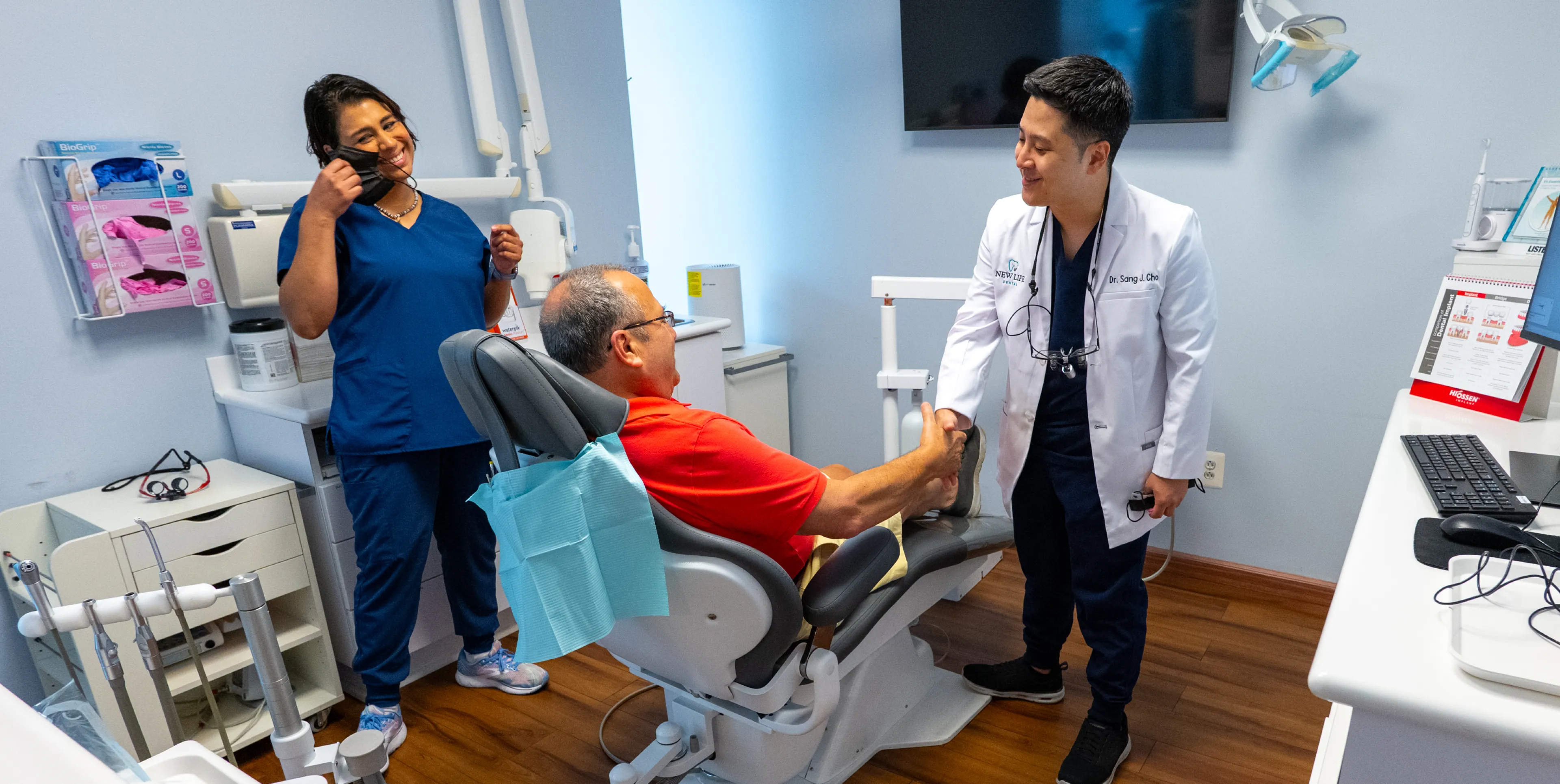 Dentist shaking hands with patient in dental chair while assistant removes her mask, smiling.
