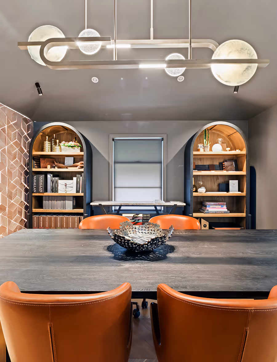 Modern dining room with a dark wooden table, four orange leather chairs, a decorative metal bowl, arched wooden bookshelves, and contemporary ceiling light fixtures.