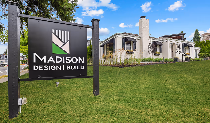Black Madison Design Build sign on green lawn in front of modern white building with awnings.