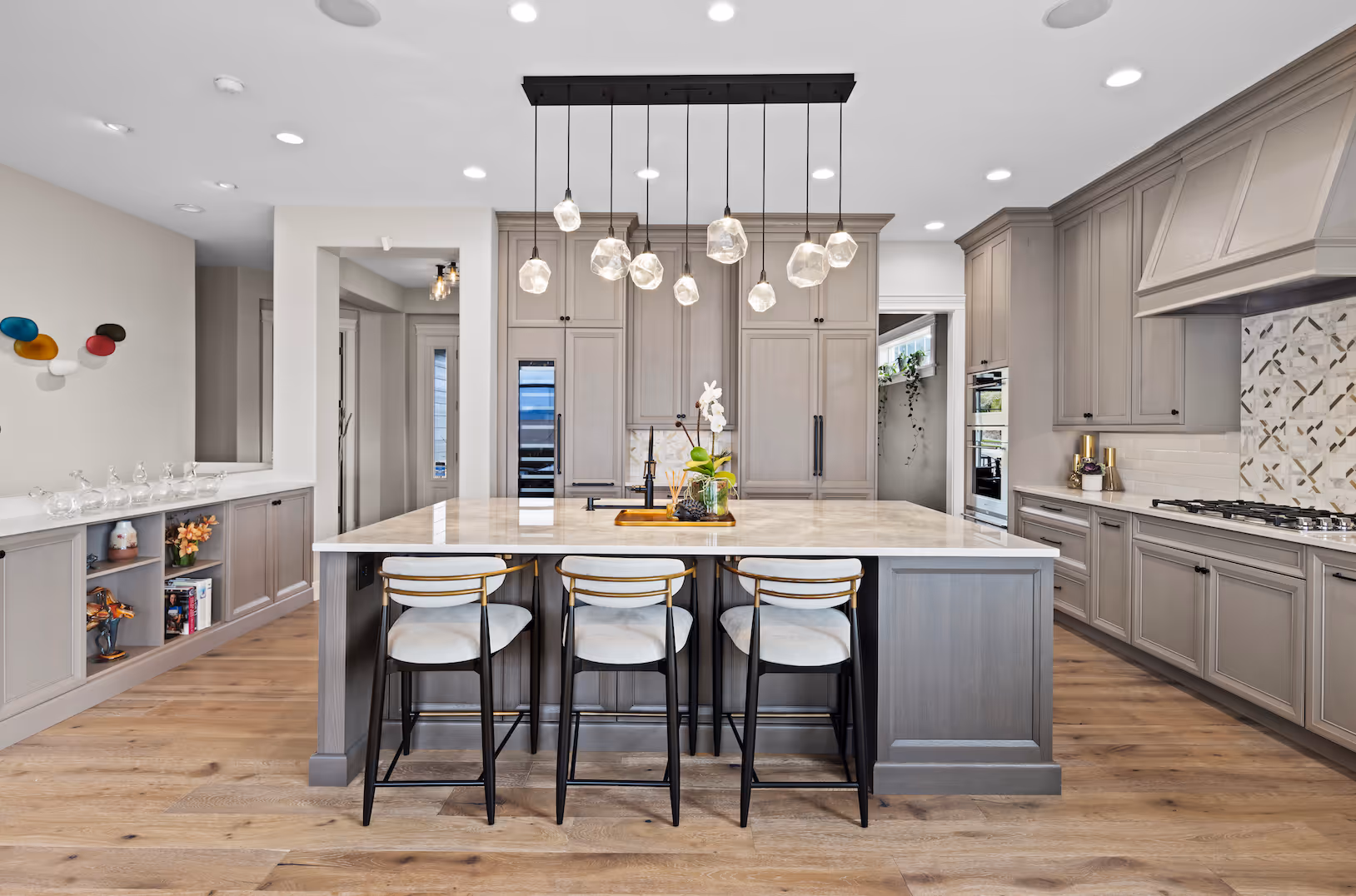 Modern kitchen with marble island, three stools, gray cabinetry, hanging lights, and wood floor.